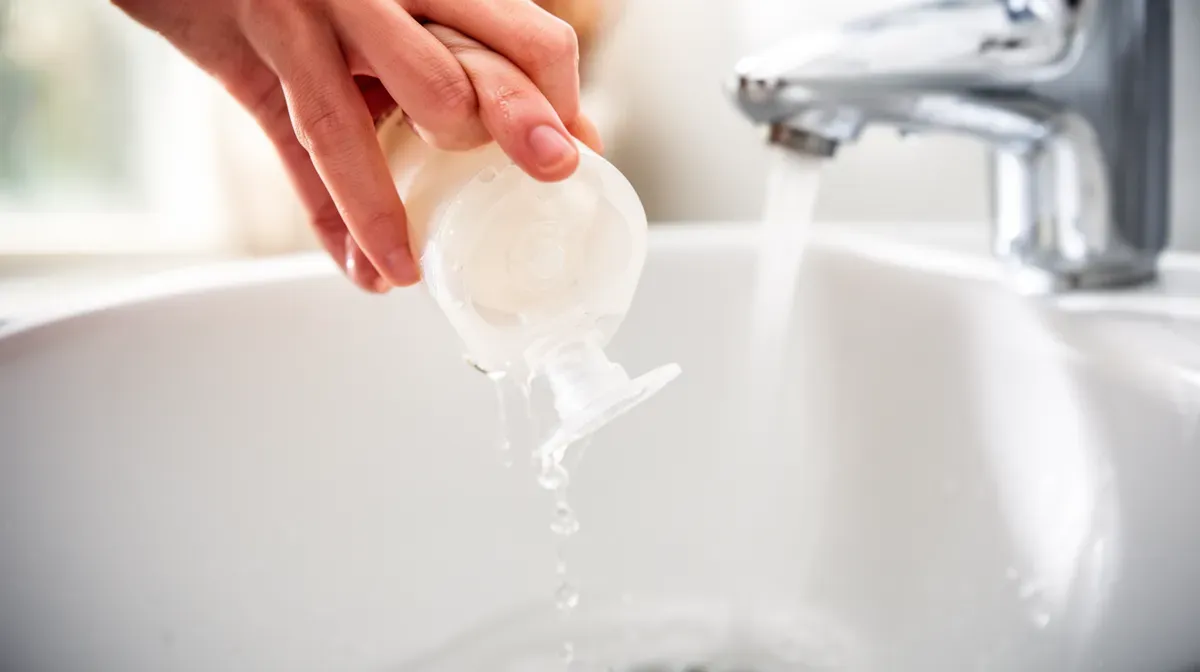 Gros plan d'une main tenant une bouteille de shampooing à l'envers au-dessus d'un évier avec de l'eau qui coule